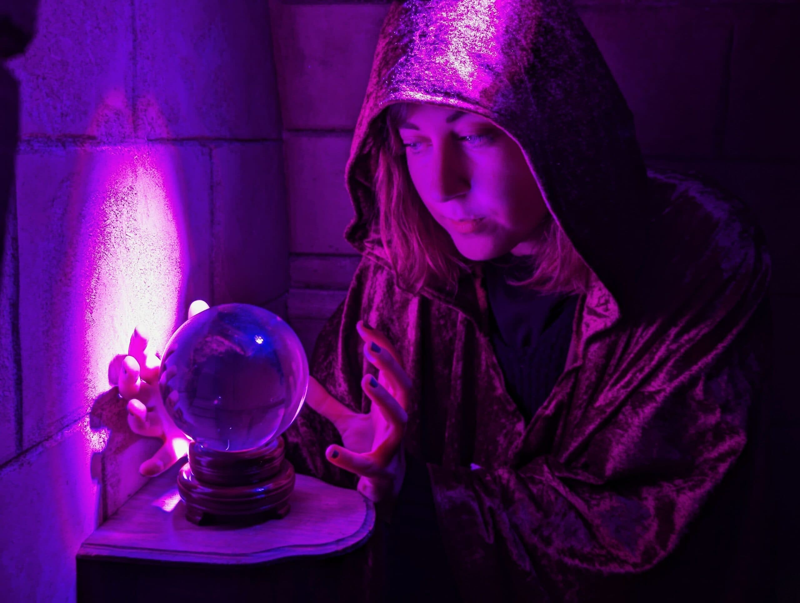 Jeune femme en cape de velours observant une boule de cristal dans une lumière violette mystérieuse, dans un décor médiéval.
